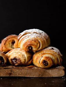 Pain Au Chocolat – được mệnh danh người anh em của bánh Croissant Pain au chocolat hay còn được gọi là bánh mì socola – 1 trong 7 món ngon Paris dành cho người hảo ngọt. Không ai biết nguồn gốc cụ thể của chiếc bánh này, nhưng tất cả đều chắc chắn rằng việc cho chocolate vào công thức làm bánh này là “một tai nạn tuyệt vời”. Lớp vỏ giòn xốp thơm ngậy, lại có thêm hương vị socola đắng đắng ngọt ngọt nồng nàn bên trong, khiến ai đã nếm một lần đều nhớ mãi không quên. Có khả năng nói người Pháp chính là bậc thầy trong việc biến những món ăn tưởng chừng giản đơn trở thành hấp dẫn và xinh xắn hơn rất nhiều. Và bánh Pain Au Chocolat chính là một trong những số đó. Bánh được làm từ bột mì, sữa tươi, bơ, trứng, ngặt nghèo lớp phủ chocolate vô cùng đơn giản tuy nhiên lại đẹp mắt và mang lại hương vị ngọt ngào khó cưỡng. nếu như là một người hảo ngọt thì bạn không nên bỏ qua loại bánh này đâu. Nguyên liệu làm bánh: Phần vỏ bánh: 60 gram bơ nhạt, để ở nhiệt độ phòng 500 gram bột mì đa dụng 50 gram đường 2 thìa cà phê muối 9.3 gram men instant 360ml sữa bò nguyên kem 115 gram socola đắng vừa, thái nhỏ Đường bột trang trí (tùy chọn) Phần bơ: 345 gram bơ nhạt, để ở nhiệt độ phòng 16 gram bột mì đa dụng Phần trứng phết lên Pain au Chocolat: 1 quả trứng 30ml sữa bò nguyên kem Cách làm gồm các bước sau đây: Bước 1: Chia 60 gram bơ ra làm 4 phần, cho vào tô cùng bột mì, đường, muối và men. Trộn đều bằng máy trộn ở tốc độ trung bình thấp. Đổ từ từ 360ml sữa vào hỗn hợp. Khi đã cho hết sữa vào, tăng tốc độ đánh lên trung bình hoặc nhồi bằng tay trong tối thiểu 5 phút. Khi quan sát thấy bột gần như tách khỏi thành tô và đàn hồi lại khi nhấn ngón tay vào là thành công. Bước 2: Phủ bột đều lên tay và mặt khay, lấy bột ra khỏi tô và nhồi thành hình tròn. Sau đó, bọc lại bằng màng bọc thực phẩm hoặc giấy bạc. Cho vào ngăn mát tủ lạnh và để bột nghỉ trong 30 phút. Bước 3: Lấy bột ra khỏi tủ lạnh và dàn phẳng bột bằng tay, sao cho phần bột thành hình tứ giác có kích thước 25x35cm. Đừng lo lắng nếu lúc này bột vẫn chưa lạnh hẳn và có cảm giác như nhào đất nặn mềm. Bước 4: Bọc lại màng bọc thực phẩm hoặc giấy bạc lên phần bột đã dàn phẳng, chú ý giữ nguyên kích thước. Để bột nghỉ trong ngăn mát tủ lạnh ít nhất 4 tiếng hoặc để qua đêm. Bước 5: Đánh đều bơ cùng bột mì đến khi hỗn hợp đều và mịn. Bỏ lên khay nướng bằng silicon để có thể dễ dàng tách ra khi cần thiết. Dùng thìa hoặc cây vét bột dàn phẳng hỗn hợp ra thành hình tứ giác kích thước 17x25cm. Cho khay bơ vào tủ lạnh trong 30 phút. Bước 6: Mang phần bột ở bước 4 và phần bơ ở bước 5 ra ngoài, để lên mặt bếp đã được phủ bột lên. Đặt phần bơ lên trên chính giữa phần bột, phủ 2 cạnh bột thừa ra lên trên và nắn để bọc hoàn toàn phần bơ vào bên trong. Sau đó, cán bột thành tứ giác kích thước 25x50cm. Khi cán, bột sẽ rất lạnh và khó thành hình tứ giác, hãy kiên nhẫn đến khi bột thành hình chính xác. Sau đó, dùng mắt chia bột thành 3 phần đều nhau rồi gấp lại. Đây là lần thứ nhất. Bước 7: Nếu lúc này bột trở nên quá ấm, cho vào khay và bọc lại bằng màng bọc thực phẩm và để bột nghỉ 30 phút trong ngăn mát tủ lạnh. Bước 8: Lấy bột ra khỏi tủ lạnh, cán bột lại thành kích thước 25x50cm như ban đầu. Sau đó, dùng mắt chia bột thành 3 phần đều nhau rồi gấp lại. Đây là lần thứ hai. Cho bột vào khay và bọc lại bằng màng bọc thực phẩm và để bột nghỉ 30 phút trong ngăn mát tủ lạnh. Bước 9: Lấy bột ra khỏi tủ lạnh, cán bột lại thành kích thước 25x50cm như ban đầu. Sau đó, dùng mắt chia bột thành 3 phần đều nhau rồi gấp lại. Bước 10: Đặt phần bột đang gấp lại lên khay nướng silicon, bọc kín lại bằng màng bọc thực phẩm. Để bột nghỉ trong ngăn mát tủ lạnh ít nhất 4 tiếng hoặc để qua đêm. Bước 11: Lấy bột ra khỏi tủ lạnh, cán thành hình tứ giác kích thước 20x50cm. Cắt dọc thành 2 phần đều nhau kích thước 20x25cm. Tiếp tục cắt ngang thành 3 phần đều nhau, để có 8 phần bột kích thước 10x12cm. Bước 12: Cắt đôi tiếp để có 16 phần bột hình tứ giác kích thước 5x12cm. Cán bột nhẹ nhàng để kéo dài ra thành 5x20cm. Cho socola vụn vào giữa, cuộn chặt và hướng phần mép bột xuống dưới. Tiếp tục đến khi hết 16 cái bánh socola. Cho bánh vào 2 khay nướng lót giấy nến, phủ màng bọc thực phẩm lên trên và để bột nghỉ khoảng 1 tiếng ở nhiệt độ thường. Sau đó, để vào ngăn mát tủ lạnh trong ít nhất 1 tiếng, tối đa 12 tiếng. Bước 13: Làm nóng lò trước ở 204°C. Trộn đều phần trứng phủ. Lấy bánh ra khỏi tủ lạnh, quét nhẹ phần trứng lên trên và cho vào lò nướng. Bước 14: Nướng khoảng 20 phút đến khi bánh vàng ruộm. Sau khoảng 10 phút đầu tiên, mở lò ra và xoay ngược khay nướng bánh lại, nếu quan sát thấy bánh chín quá nhanh, hạ nhiệt độ xuống 190°C. Bước 15: Sau khi lấy bánh ra khỏi lò, để trên giá vài phút để bánh nguội bớt. Bánh ngon nhất khi ăn luôn trong ngày. Tuy nhiên, có thể để bánh 2-3 ngày ở ngoài hoặc 1 tuần trong ngăn mát tủ lạnh. Chúc các bạn hoàn thành món bánh này để có thể tặng cho bạn bè hoặc thay đổi món ăn vặt cho gia đình bạn. Bạn có thể theo dõi chúng tôi để có thể làm ra nhiều món ăn khác nhau nhé! Đối với các cơ sở - doanh nghiệp quan tâm đến các loại máy đóng gói có thể liên lạc với Máy đóng gói An Thành - chúng tôi để được tư vấn, hỗ trợ để tìm cho bạn chiếc máy phù hợp sản phẩm mà bạn đang đóng gói. CÔNG TY TNHH SẢN XUẤT – THƯƠNG MẠI – DỊCH VỤ – AN THÀNH Địa chỉ: 47/80 Ao Đôi, Khu Phố 10, Phường Bình Trị Đông A, Quận Bình Tân, TP.HCM Email: anthanhsale01@gmail.com Hotline (zalo) :  0903103922 – 0906312325 Website: https://www.packvn.com/ – https://maydonggoi.vip/ Facebook: https://www.facebook.com/maydonggoianthanh Pinterest: https://www.pinterest.com/donggoianthanh/_saved/ Instagram: https://www.instagram.com/maydonggoianthanh/ Twitter:https://twitter.com/donggoianthanh Linkedin: https://www.linkedin.com/in/may-dong-goi-bao-bi-an-thanh/ Youtube: Máy đóng gói An Thành – YouTube.