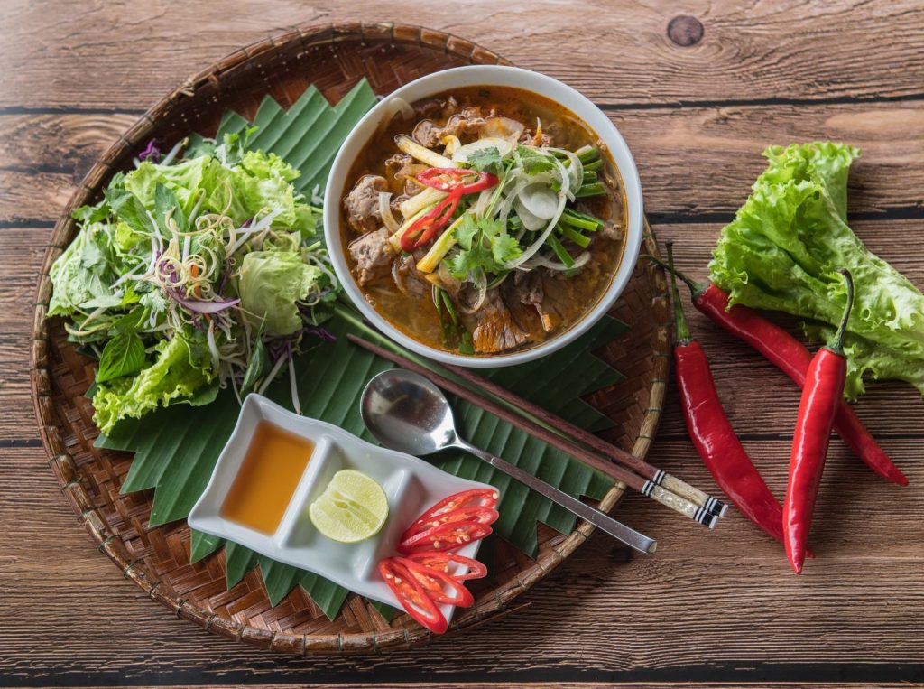 Bún bò Huế là một trong những đặc sản của xứ Huế, mặc dù món bún này phổ biến trên cả ba miền ở Việt Nam và ở nước ngoài. Tại Huế, món này được gọi đơn giản là "bún bò" hoặc gọi cụ thể hơn là "bún bò giò heo". Các địa phương khác gọi là "bún bò Huế", "bún bò gốc Huế" để chỉ xuất xứ của món ăn này. Món ăn có nguyên liệu chính là bún, thịt bắp bò, giò heo, cùng nước dùng có màu đỏ đặc trưng và vị sả và ruốc. Đôi khi tô bún còn được thêm vào thịt bò tái, chả cua, .... các loại nguyên liệu khác tùy theo sở thích của người nấu và tùy thuộc vào mỗi vùng miền khác nhau cách nêm nếm sẽ khác nhau để cho phù hợp cách ăn nơi đó. Ở xứ Huế, người ta có cách nêm nước dùng rất riêng biệt như thường nêm vào một ít mắm ruốc, góp phần làm nên hương vị rất riêng của nồi bún bò Huế. Sau khi xương bò được hầm chín tới, người ta thường thêm vào một ít chả heo hay chả cua được quết nhuyễn. Thịt bò có thể được cắt mỏng, nhúng vào nước dùng đang sôi trước khi cho vào tô bún (gọi là thịt bò tái). Người ta cũng thường cho thêm một ít ớt bột và gia vị vào tô bún rồi ăn với rau sống gồm giá, rau thơm, xà lách, rau cải con, bắp chuối cắt nhỏ. Nguyên liệu nấu bún bò Huế, ta có gì: Sợi bún bò 500gr bắp bò 500gr xương heo hoặc xương bò Chả Huế 3 muỗng mắm ruốc Chả cua Sả, gừng, tỏi, hành tím, ớt bột Thảo quả, hoa hồi, thanh quế 1 củ hành tây Hạt màu điều Chanh, ớt, hành lá, rau răm Các loại rau sống ăn kèm: Giá, xà lách, Húng quế, tía tô, bắp chuối bào, rau muống bào,… Gia vị: Hạt nêm, muối, nước mắm, bột ngọt, đường, dầu ăn,…. Cách nấu bún bò Huế: Bước 1: Sơ chế nguyên liệu – Rửa sạch xương, rồi trụng sơ qua với nước sôi để loại bỏ chất dơ, nồi nước dùng sẽ trong hơn sau khi nấu. – Gừng, hành tím làm sạch vỏ, sau đó cho vào trong giấy bạc với hoa hồi và quế, đem đi nướng ở trong lò 7 phút ở nhiệt độ 180 độ C. – Sả rửa sạch, đập dập rồi cắt thành từng khúc khoảng 5cm. – Hành tây lột vỏ, rửa sạch, rồi cắt thành từng lát mỏng, sau đó cho vào trong tô nước đá lạnh, ngâm trong 5 – 10 phút để giảm bớt mùi hăng của hành. – Rau răm, hành lá rửa sạch, với rau răm thì cắt nhỏ, hành lá thì cắt phần đầu hành để riêng, phần lá thì cắt nhỏ. – Chanh và ớt rửa sạch, cắt theo sở thích để ăn kèm với bún. – Các loại rau ăn kèm, giá nhặt và rửa sạch – Hòa tan mắm ruốc vào 1 chén nước để qua 1 bên – Huyết (hay còn gọi là tiết): Thường thì huyết bò sẽ được sử dụng nhiều để đảm bảo sự thống nhất của hương vị. – Chả cua: nếu có thể tự làm ra được là tốt nhất, do cách làm đơn giản, không quá khó. Nhưng nếu điều kiện không cho phép, nên mua tại nơi uy tín để đảm bảo chất lượng. Chả cua là thành phần tạo nên tô bún bò ngon chính hiệu Huế. Có thể sử dụng thêm giò heo hoặc giò bò để tăng độ đa dạng và kích thích của món ăn. – Thịt bò mua về thì rửa sạch. Bắp bò và chân giò: Lựa chọn đồ tươi, đảm bảo chất lượng thịt. Bắp bò có thể sử dụng luộc chung với nước dùng để tạo ra độ ngọt cho nước dùng. Chân giò nên luộc riêng, để tránh làm đục nước. – Bún: thường là bún sợi to, đảm bảo nguồn cung cấp để tránh những nơi cung cấp bún chứa hàn the hoặc huỳnh quang. Bước 2: Làm màu điều – Cho một ít dầu ăn vào nồi, sau đó cho hạt màu điều vào, để hạt màu điều ra hết màu thì tắt bếp. Lọc bỏ phần xác, rồi cho phần dầu màu điều vào trong chén. Bước 3: Làm sa tế ăn bún bò Huế – Trong thời gian chờ nước dùng thì làm sa tế. Băm nhuyễn 1 củ hành khô, nửa củ tỏi và 2 cây sả. Bắc chảo lên bếp, khi chảo nóng thì cho dầu ăn vào phi thơm sả, hành và tỏi đến khi vàng thì đổ ớt bột vào đảo qua lại và tắt bếp. Lưu ý: Không để lửa to quá sẽ khiến ớt bị cháy ăn sẽ bị đắng. Bước 4: Nấu nước dùng bún bò Huế – Cho xương vào trong nồi cùng với 2 lít nước, sau đó tiến hành hầm xương ở lửa nhỏ trong 1 tiếng để xương ra hết chất ngọt. Sau 30 phút, vớt bỏ bọt, rồi cho hết phần gừng, hành tím, hoa hồi, quế, thảo quả, sả và thịt bò vào, đậy nắp lại rồi tiếp tục nấu. – Sau đó vớt gừng, hành tím, hoa hồi, quế, thảo quả và sả ra. Nấu thêm 15 phút, thì vớt thịt bò ra để nguội sau đó cắt miếng vừa ăn. Cho chén mắm ruốc đã pha vào nồi nước, nêm nếm với gia vị cho vừa ăn. Lưu ý: Hầm xương nhỏ lửa, trong quá trình ninh, nếu thêm nước nên thêm nước nóng, để đảm bảo nước hầm giữ được độ ngọt của xương. Để nước dùng ngon nhất, bạn nên sử dụng nồi hầm xương trong bộ nồi nấu phở bằng điện sẽ giữ được độ ngon, ngọt của xương, chuẩn vị bún bò Huế. – Mắm ruốc cho vào nước lạnh khuấy đều và tan, sau đó đợi phần bã lắng xuống, lấy nước cho vào nồi nước dùng để có được hương vị đặc trưng của bún bò Huế. – Để nước dùng thơm và có màu đẹp: Làm nóng 2 muỗng canh dầu, phi thơm 1,5 muỗng canh sả băm, 1 muỗng canh tỏi băm. Khi sả và tỏi săn lại, thơm thì cho dầu màu điều đã làm vào. Tắt bếp. Sau đó cho vào nồi nước dùng. – Cho bún ra tô, thịt bò cắt lát mỏng, chả Huế. Múc nước dùng cho vào tô bún, rắc lên một ít hành tây, rau răm và hành lá vào. Bún bò ăn kèm các loại rau. Bí quyết làm nồi nước lèo bún bò Huế đậm đà chuẩn vị Huế bạn cần biết: Đầu tiên là lựa chọn thịt bò và chân giò tươi: Thịt bò phù hợp cho món bún bò Huế này chính là bắp bò, nếu được thì chọn loại bắp hoa thì mới thơm mới ngon được. Chân giò nên chọn chân giò phía trước, thường thì nó sẽ có thịt săn ăn sẽ thơm ngon hơn. Nếu được bạn cũng có thể nhờ người bán lựa chọn đúng loại thịt bò cũng như tham khảo thêm cách lựa thịt bò phù hợp. Mua thêm một ít xương bò để ninh cùng trong nồi nước lèo bún bò Huế. Sơ chế thịt bò: Chân giò sau khi mua về thì cạo thật sạch lông, cắt hết móng đi và rửa sạch lại. Chặt chân giò thành những khoanh tròn vừa phải. Cho chân giò và xương bò vào nồi nước sôi luộc đến khi nước sôi trở lại thì chắt bỏ bớt. Sau đó rửa lại chân giò cùng xương bò cho sạch các bọt bẩn. Còn về phần thịt bò sau khi mua về bạn rửa sạch và cắt lát mỏng vừa ăn. Bạn cũng nên lưu ý chọn thịt để làm bún bò Huế là thịt bắp bò, khi ăn sẽ ngon hơn nhờ phần gân kèm theo từng sới thịt săn chắc. Hiện đang vào mùa mưa và tình hình dịch bệnh đang hoành hành mạnh cho nên ta cần bảo vệ tốt sức khỏe của bản thân và gia đình mình. Vậy ăn gì vào mùa mưa bão để tốt cho sức khỏe? Ăn nhiều trái cây: Vào tiết giao mùa mưa bão, thời tiết thay đổi thất thường thì bạn càng cần ăn đủ lượng trái cây cần thiết để có sức khoẻ tốt, đặc biệt là các loại trái cây giàu chất chống oxy hoá, giàu vitamin và chất khoáng có tác dụng hỗ trợ và tăng cường hệ miễn dịch. Bổ sung rau củ quả cho thực đơn hằng ngày: Ngoài tăng cường ăn đủ các loại rau củ, bạn nên tập trung ăn các loại rau họ đắng. Vì rau họ đắng được xem như loại thực phẩm cần thiết khi giao mùa từ nóng sang lạnh. Chúng giúp cơ thể tránh khỏi nguy cơ bị nhiễm trùng da hay dị ứng - hai loại bệnh phổ biến khi mưa bão. Các gia vị nào ta cần bổ sung trong bữa ăn: Tỏi - Loại gia vị rất giàu chất chống oxy hóa nên có tác dụng hỗ trợ và tăng cường hệ miễn dịch hiệu quả. Ngoài ra, hệ tiêu hóa của bạn cũng được giảm bớt áp lực nhờ khả năng thúc đẩy quá trình trao đổi chất của tỏi. Đặc biệt, khi trời chúng ta có xu hướng ăn đồ nướng, cay nóng nhiều hơn khiến dạ dày chịu áp lực lớn. Gừng - Không chỉ dùng làm gia vị cho bữa ăn, gừng cũng có thể được dùng pha trà cho người vừa đi mưa về giúp cải thiện hệ miễn dịch, làm ấm cơ thể và thúc đẩy quá trình trao đổi chất hiệu quả. Nghệ - Giúp bạn kiểm soát được đường huyết cơ thể và giảm nguy cơ mắc các bệnh do gió mùa. Hạt tiêu đen - Gia vị có đặc tính giúp cơ thể giải độc khi bị các bệnh như ho, cảm lạnh, cảm cúm, đau cơ hay những dạng bệnh hô hấp khi mưa gió. Do vậy mà hạt tiêu đen là một loại gia vị mùa mưa bão nên ăn. Nước là thứ không thể thiếu: Mùa mưa bão không khí ẩm lạnh thường khiến bạn quên cảm giác khát nước, từ đó cơ thể có nguy cơ bị thiếu hụt nước trầm trọng và làm hệ miễn dịch suy yếu, tăng nguy cơ mắc bệnh về huyết áp, nhiễm trùng hoặc bệnh truyền nhiễm. Để đảm bảo sức khỏe tốt, bạn cần đảm bảo lượng nước cho cơ thể hằng ngày. Hãy uống ít nhất 8 ly nước với dung tích 150ml/ngày. Giữ vệ sinh an toàn thực phẩm vào mùa mưa: Với những vùng ít bị ảnh hưởng do bão lũ, mưa nhiều vẫn có thể gây ra các vấn đề về sức khỏe cũng như an toàn thực phẩm. Bạn nên: Giữ bếp khô ráo, vệ sinh và lau chùi thường xuyên mặt bếp, dụng cụ làm bếp và cả các loại khăn lau Cất trữ thực phẩm trong hộp kín. Chọn các loại hộp không thấm nước và đảm bảo nắp đậy kín, bảo quản tốt thực phẩm. Để thực phẩm ở nơi cao, thoáng mát. Luôn ăn chín, uống sôi. Ưu tiên chọn thực phẩm tươi, rửa sạch và sử dụng ngay. Thức ăn cần được nấu chín ở nhiệt độ cao, uống nước đun sôi để nguội. Hạn chế dùng ngay thực phẩm đã bảo quản lâu ngày trong tủ lạnh. Nếu bạn muốn ăn thực phẩm đã nấu chín và cất tủ lạnh trước đó, hãy đun nóng dần cho đến khi sôi trở lại rồi mới sử dụng. Tình hình thời tiết luôn diễn ra bất ngờ, dù được dự báo trước nhưng chúng ta thường ít lường được mức độ nghiêm trọng và những thiệt hại thực tế ảnh hưởng đến đời sống, tinh thần và sức khỏe của mỗi người. CÔNG TY TNHH SẢN XUẤT – THƯƠNG MẠI – DỊCH VỤ – AN THÀNH Địa chỉ: 47/80 Ao Đôi, Khu Phố 10, Phường Bình Trị Đông A, Quận Bình Tân, TP.HCM Email: anthanhsale01@gmail.com Hotline (zalo) :  0903103922 – 0906312325 Website: https://www.packvn.com/ – https://maydonggoi.vip/ Facebook: https://www.facebook.com/maydonggoianthanh Pinterest: https://www.pinterest.com/donggoianthanh/_saved/ Instagram: https://www.instagram.com/maydonggoianthanh/ Twitter:https://twitter.com/donggoianthanh Linkedin: https://www.linkedin.com/in/may-dong-goi-bao-bi-an-thanh/ Youtube: Máy đóng gói An Thành – YouTube.