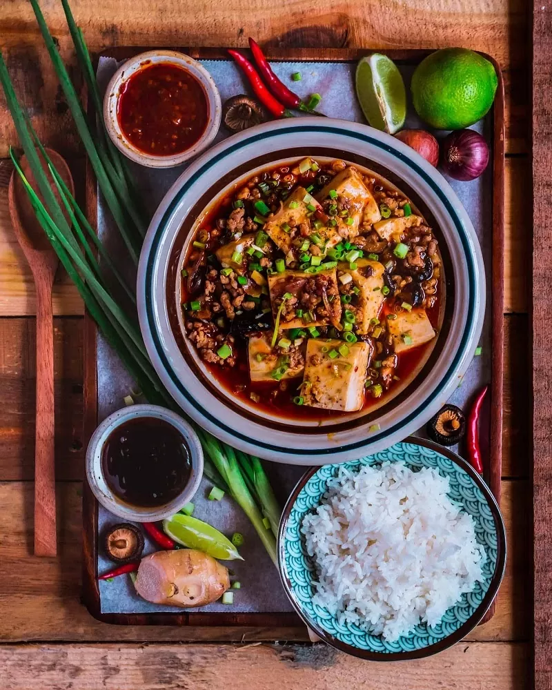 Đậu hũ Tứ Xuyên làm món ăn đặc sản của vùng Tứ Xuyên - Trung Hoa. Mang hương vị cay đặc trưng , cảm giác bạn sẽ không bao giờ quên nếu bạn đã từng thưởng thức. Sự kết hợp giữa miếng đậu hũ trắng mềm, thịt xay và ớt cay riêng biệt của vùng Tứ Xuyên đã tạo nên một món ăn mang cả hương vị lẫn màu sắc vô cùng bắt mắt Vậy theo bạn, món ăn đậu hũ Tứ Xuyên - Mapo đã được ra đời như thế nào? Món ăn đơn giản từ trong nguyên liệu cho đến cách chế biến này lại trở thành niềm tự hào của người Trung Hoa. Tuy đơn giản nhưng lại chứa đựng hương vị thơm ngon và cả một lịch sử hình thành, phát triển thú vị. Đậu hũ Tứ Xuyên thực chất có tên gốc là Mapo. Trong tiếng Trung Quốc, “Mapo” là từ ghép có ý chỉ một người phụ nữ lớn tuổi và mặt rỗ. Sở dĩ món ăn có cái tên này là bởi nó gắn liền với truyền thuyết về nhà hàng của bà Chen: Tứ Xuyên xưa kia có một hàng cơm nhỏ do bà Chen Mapo làm chủ. Không ai rõ tên thực của bà là gì, chỉ biết bà từ nhỏ do bị bệnh đã để lại sẹo rỗ trên mặt, nên người ta thường gọi là “Bà Chen mặt rỗ” – tức Chen Mapo. Để xử lý số lượng đậu hũ dư thừa trong kho thực phẩm, Chen Mapo đã nghĩ ra cách kết hợp nó với các nguyên liệu khác nhằm đổi mới món ăn, đưa ra một công thức hoàn toàn mới. Bà đã trộn đậu với thịt heo băm nhỏ cùng rất nhiều gia vị khác nhau, xào chung trên một chiếc chảo lớn. Công thức này mau chóng đem lại tiếng tăm cho nhà hàng của Chen Mapo. Sau này, khi món ăn có mặt tại nhiều nhà hàng lớn, người ta đã đổi tên thành nơi sản sinh ra nó – Tứ Xuyên. Món đậu hũ Tứ Xuyên là sự tổng hòa của nhiều nguyên liệu để cho ra 7 loại hương vị hấp dẫn và kích thích mạnh đến vị giác người dùng đó là: bùi – cay – nóng – tươi – mềm – thơm – giòn. Hãy cùng vào bếp và thực hiện món ăn này với các bước hướng dẫn cùng chúng tôi để cảm nhận những tinh túy trong ẩm thực Trung Hoa nhé!