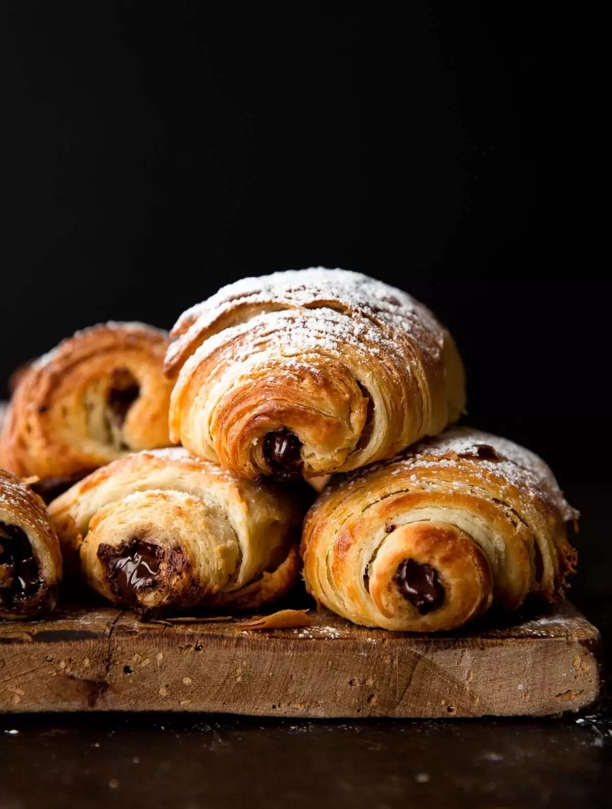Pain Au Chocolat – được mệnh danh người anh em của bánh Croissant Pain au chocolat hay còn được gọi là bánh mì socola – 1 trong 7 món ngon Paris dành cho người hảo ngọt. Không ai biết nguồn gốc cụ thể của chiếc bánh này, nhưng tất cả đều chắc chắn rằng việc cho chocolate vào công thức làm bánh này là “một tai nạn tuyệt vời”. Lớp vỏ giòn xốp thơm ngậy, lại có thêm hương vị socola đắng đắng ngọt ngọt nồng nàn bên trong, khiến ai đã nếm một lần đều nhớ mãi không quên. Có khả năng nói người Pháp chính là bậc thầy trong việc biến những món ăn tưởng chừng giản đơn trở thành hấp dẫn và xinh xắn hơn rất nhiều. Và bánh Pain Au Chocolat chính là một trong những số đó. Bánh được làm từ bột mì, sữa tươi, bơ, trứng, ngặt nghèo lớp phủ chocolate vô cùng đơn giản tuy nhiên lại đẹp mắt và mang lại hương vị ngọt ngào khó cưỡng. nếu như là một người hảo ngọt thì bạn không nên bỏ qua loại bánh này đâu. Nguyên liệu làm bánh: Phần vỏ bánh: 60 gram bơ nhạt, để ở nhiệt độ phòng 500 gram bột mì đa dụng 50 gram đường 2 thìa cà phê muối 9.3 gram men instant 360ml sữa bò nguyên kem 115 gram socola đắng vừa, thái nhỏ Đường bột trang trí (tùy chọn) Phần bơ: 345 gram bơ nhạt, để ở nhiệt độ phòng 16 gram bột mì đa dụng Phần trứng phết lên Pain au Chocolat: 1 quả trứng 30ml sữa bò nguyên kem Cách làm gồm các bước sau đây: Bước 1: Chia 60 gram bơ ra làm 4 phần, cho vào tô cùng bột mì, đường, muối và men. Trộn đều bằng máy trộn ở tốc độ trung bình thấp. Đổ từ từ 360ml sữa vào hỗn hợp. Khi đã cho hết sữa vào, tăng tốc độ đánh lên trung bình hoặc nhồi bằng tay trong tối thiểu 5 phút. Khi quan sát thấy bột gần như tách khỏi thành tô và đàn hồi lại khi nhấn ngón tay vào là thành công. Bước 2: Phủ bột đều lên tay và mặt khay, lấy bột ra khỏi tô và nhồi thành hình tròn. Sau đó, bọc lại bằng màng bọc thực phẩm hoặc giấy bạc. Cho vào ngăn mát tủ lạnh và để bột nghỉ trong 30 phút. Bước 3: Lấy bột ra khỏi tủ lạnh và dàn phẳng bột bằng tay, sao cho phần bột thành hình tứ giác có kích thước 25x35cm. Đừng lo lắng nếu lúc này bột vẫn chưa lạnh hẳn và có cảm giác như nhào đất nặn mềm. Bước 4: Bọc lại màng bọc thực phẩm hoặc giấy bạc lên phần bột đã dàn phẳng, chú ý giữ nguyên kích thước. Để bột nghỉ trong ngăn mát tủ lạnh ít nhất 4 tiếng hoặc để qua đêm. Bước 5: Đánh đều bơ cùng bột mì đến khi hỗn hợp đều và mịn. Bỏ lên khay nướng bằng silicon để có thể dễ dàng tách ra khi cần thiết. Dùng thìa hoặc cây vét bột dàn phẳng hỗn hợp ra thành hình tứ giác kích thước 17x25cm. Cho khay bơ vào tủ lạnh trong 30 phút. Bước 6: Mang phần bột ở bước 4 và phần bơ ở bước 5 ra ngoài, để lên mặt bếp đã được phủ bột lên. Đặt phần bơ lên trên chính giữa phần bột, phủ 2 cạnh bột thừa ra lên trên và nắn để bọc hoàn toàn phần bơ vào bên trong. Sau đó, cán bột thành tứ giác kích thước 25x50cm. Khi cán, bột sẽ rất lạnh và khó thành hình tứ giác, hãy kiên nhẫn đến khi bột thành hình chính xác. Sau đó, dùng mắt chia bột thành 3 phần đều nhau rồi gấp lại. Đây là lần thứ nhất. Bước 7: Nếu lúc này bột trở nên quá ấm, cho vào khay và bọc lại bằng màng bọc thực phẩm và để bột nghỉ 30 phút trong ngăn mát tủ lạnh. Bước 8: Lấy bột ra khỏi tủ lạnh, cán bột lại thành kích thước 25x50cm như ban đầu. Sau đó, dùng mắt chia bột thành 3 phần đều nhau rồi gấp lại. Đây là lần thứ hai. Cho bột vào khay và bọc lại bằng màng bọc thực phẩm và để bột nghỉ 30 phút trong ngăn mát tủ lạnh. Bước 9: Lấy bột ra khỏi tủ lạnh, cán bột lại thành kích thước 25x50cm như ban đầu. Sau đó, dùng mắt chia bột thành 3 phần đều nhau rồi gấp lại. Bước 10: Đặt phần bột đang gấp lại lên khay nướng silicon, bọc kín lại bằng màng bọc thực phẩm. Để bột nghỉ trong ngăn mát tủ lạnh ít nhất 4 tiếng hoặc để qua đêm. Bước 11: Lấy bột ra khỏi tủ lạnh, cán thành hình tứ giác kích thước 20x50cm. Cắt dọc thành 2 phần đều nhau kích thước 20x25cm. Tiếp tục cắt ngang thành 3 phần đều nhau, để có 8 phần bột kích thước 10x12cm. Bước 12: Cắt đôi tiếp để có 16 phần bột hình tứ giác kích thước 5x12cm. Cán bột nhẹ nhàng để kéo dài ra thành 5x20cm. Cho socola vụn vào giữa, cuộn chặt và hướng phần mép bột xuống dưới. Tiếp tục đến khi hết 16 cái bánh socola. Cho bánh vào 2 khay nướng lót giấy nến, phủ màng bọc thực phẩm lên trên và để bột nghỉ khoảng 1 tiếng ở nhiệt độ thường. Sau đó, để vào ngăn mát tủ lạnh trong ít nhất 1 tiếng, tối đa 12 tiếng. Bước 13: Làm nóng lò trước ở 204°C. Trộn đều phần trứng phủ. Lấy bánh ra khỏi tủ lạnh, quét nhẹ phần trứng lên trên và cho vào lò nướng. Bước 14: Nướng khoảng 20 phút đến khi bánh vàng ruộm. Sau khoảng 10 phút đầu tiên, mở lò ra và xoay ngược khay nướng bánh lại, nếu quan sát thấy bánh chín quá nhanh, hạ nhiệt độ xuống 190°C. Bước 15: Sau khi lấy bánh ra khỏi lò, để trên giá vài phút để bánh nguội bớt. Bánh ngon nhất khi ăn luôn trong ngày. Tuy nhiên, có thể để bánh 2-3 ngày ở ngoài hoặc 1 tuần trong ngăn mát tủ lạnh. Chúc các bạn hoàn thành món bánh này để có thể tặng cho bạn bè hoặc thay đổi món ăn vặt cho gia đình bạn. Bạn có thể theo dõi chúng tôi để có thể làm ra nhiều món ăn khác nhau nhé! Đối với các cơ sở - doanh nghiệp quan tâm đến các loại máy đóng gói có thể liên lạc với Máy đóng gói An Thành - chúng tôi để được tư vấn, hỗ trợ để tìm cho bạn chiếc máy phù hợp sản phẩm mà bạn đang đóng gói. CÔNG TY TNHH SẢN XUẤT – THƯƠNG MẠI – DỊCH VỤ – AN THÀNH Địa chỉ: 47/80 Ao Đôi, Khu Phố 10, Phường Bình Trị Đông A, Quận Bình Tân, TP.HCM Email: anthanhsale01@gmail.com Hotline (zalo) : 0903103922 – 0906312325 Website: https://www.packvn.com/ – https://maydonggoi.vip/ Facebook: https://www.facebook.com/maydonggoianthanh Pinterest: https://www.pinterest.com/donggoianthanh/_saved/ Instagram: https://www.instagram.com/maydonggoianthanh/ Twitter:https://twitter.com/donggoianthanh Linkedin: https://www.linkedin.com/in/may-dong-goi-bao-bi-an-thanh/ Youtube: Máy đóng gói An Thành – YouTube.
