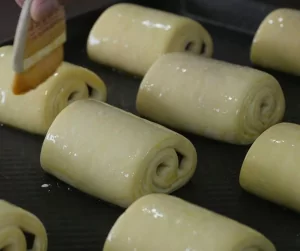 Pain Au Chocolat – được mệnh danh người anh em của bánh Croissant Pain au chocolat hay còn được gọi là bánh mì socola – 1 trong 7 món ngon Paris dành cho người hảo ngọt. Không ai biết nguồn gốc cụ thể của chiếc bánh này, nhưng tất cả đều chắc chắn rằng việc cho chocolate vào công thức làm bánh này là “một tai nạn tuyệt vời”. Lớp vỏ giòn xốp thơm ngậy, lại có thêm hương vị socola đắng đắng ngọt ngọt nồng nàn bên trong, khiến ai đã nếm một lần đều nhớ mãi không quên. Có khả năng nói người Pháp chính là bậc thầy trong việc biến những món ăn tưởng chừng giản đơn trở thành hấp dẫn và xinh xắn hơn rất nhiều. Và bánh Pain Au Chocolat chính là một trong những số đó. Bánh được làm từ bột mì, sữa tươi, bơ, trứng, ngặt nghèo lớp phủ chocolate vô cùng đơn giản tuy nhiên lại đẹp mắt và mang lại hương vị ngọt ngào khó cưỡng. nếu như là một người hảo ngọt thì bạn không nên bỏ qua loại bánh này đâu. Nguyên liệu làm bánh: Phần vỏ bánh: 60 gram bơ nhạt, để ở nhiệt độ phòng 500 gram bột mì đa dụng 50 gram đường 2 thìa cà phê muối 9.3 gram men instant 360ml sữa bò nguyên kem 115 gram socola đắng vừa, thái nhỏ Đường bột trang trí (tùy chọn) Phần bơ: 345 gram bơ nhạt, để ở nhiệt độ phòng 16 gram bột mì đa dụng Phần trứng phết lên Pain au Chocolat: 1 quả trứng 30ml sữa bò nguyên kem Cách làm gồm các bước sau đây: Bước 1: Chia 60 gram bơ ra làm 4 phần, cho vào tô cùng bột mì, đường, muối và men. Trộn đều bằng máy trộn ở tốc độ trung bình thấp. Đổ từ từ 360ml sữa vào hỗn hợp. Khi đã cho hết sữa vào, tăng tốc độ đánh lên trung bình hoặc nhồi bằng tay trong tối thiểu 5 phút. Khi quan sát thấy bột gần như tách khỏi thành tô và đàn hồi lại khi nhấn ngón tay vào là thành công. Bước 2: Phủ bột đều lên tay và mặt khay, lấy bột ra khỏi tô và nhồi thành hình tròn. Sau đó, bọc lại bằng màng bọc thực phẩm hoặc giấy bạc. Cho vào ngăn mát tủ lạnh và để bột nghỉ trong 30 phút. Bước 3: Lấy bột ra khỏi tủ lạnh và dàn phẳng bột bằng tay, sao cho phần bột thành hình tứ giác có kích thước 25x35cm. Đừng lo lắng nếu lúc này bột vẫn chưa lạnh hẳn và có cảm giác như nhào đất nặn mềm. Bước 4: Bọc lại màng bọc thực phẩm hoặc giấy bạc lên phần bột đã dàn phẳng, chú ý giữ nguyên kích thước. Để bột nghỉ trong ngăn mát tủ lạnh ít nhất 4 tiếng hoặc để qua đêm. Bước 5: Đánh đều bơ cùng bột mì đến khi hỗn hợp đều và mịn. Bỏ lên khay nướng bằng silicon để có thể dễ dàng tách ra khi cần thiết. Dùng thìa hoặc cây vét bột dàn phẳng hỗn hợp ra thành hình tứ giác kích thước 17x25cm. Cho khay bơ vào tủ lạnh trong 30 phút. Bước 6: Mang phần bột ở bước 4 và phần bơ ở bước 5 ra ngoài, để lên mặt bếp đã được phủ bột lên. Đặt phần bơ lên trên chính giữa phần bột, phủ 2 cạnh bột thừa ra lên trên và nắn để bọc hoàn toàn phần bơ vào bên trong. Sau đó, cán bột thành tứ giác kích thước 25x50cm. Khi cán, bột sẽ rất lạnh và khó thành hình tứ giác, hãy kiên nhẫn đến khi bột thành hình chính xác. Sau đó, dùng mắt chia bột thành 3 phần đều nhau rồi gấp lại. Đây là lần thứ nhất. Bước 7: Nếu lúc này bột trở nên quá ấm, cho vào khay và bọc lại bằng màng bọc thực phẩm và để bột nghỉ 30 phút trong ngăn mát tủ lạnh. Bước 8: Lấy bột ra khỏi tủ lạnh, cán bột lại thành kích thước 25x50cm như ban đầu. Sau đó, dùng mắt chia bột thành 3 phần đều nhau rồi gấp lại. Đây là lần thứ hai. Cho bột vào khay và bọc lại bằng màng bọc thực phẩm và để bột nghỉ 30 phút trong ngăn mát tủ lạnh. Bước 9: Lấy bột ra khỏi tủ lạnh, cán bột lại thành kích thước 25x50cm như ban đầu. Sau đó, dùng mắt chia bột thành 3 phần đều nhau rồi gấp lại. Bước 10: Đặt phần bột đang gấp lại lên khay nướng silicon, bọc kín lại bằng màng bọc thực phẩm. Để bột nghỉ trong ngăn mát tủ lạnh ít nhất 4 tiếng hoặc để qua đêm. Bước 11: Lấy bột ra khỏi tủ lạnh, cán thành hình tứ giác kích thước 20x50cm. Cắt dọc thành 2 phần đều nhau kích thước 20x25cm. Tiếp tục cắt ngang thành 3 phần đều nhau, để có 8 phần bột kích thước 10x12cm. Bước 12: Cắt đôi tiếp để có 16 phần bột hình tứ giác kích thước 5x12cm. Cán bột nhẹ nhàng để kéo dài ra thành 5x20cm. Cho socola vụn vào giữa, cuộn chặt và hướng phần mép bột xuống dưới. Tiếp tục đến khi hết 16 cái bánh socola. Cho bánh vào 2 khay nướng lót giấy nến, phủ màng bọc thực phẩm lên trên và để bột nghỉ khoảng 1 tiếng ở nhiệt độ thường. Sau đó, để vào ngăn mát tủ lạnh trong ít nhất 1 tiếng, tối đa 12 tiếng. Bước 13: Làm nóng lò trước ở 204°C. Trộn đều phần trứng phủ. Lấy bánh ra khỏi tủ lạnh, quét nhẹ phần trứng lên trên và cho vào lò nướng. Bước 14: Nướng khoảng 20 phút đến khi bánh vàng ruộm. Sau khoảng 10 phút đầu tiên, mở lò ra và xoay ngược khay nướng bánh lại, nếu quan sát thấy bánh chín quá nhanh, hạ nhiệt độ xuống 190°C. Bước 15: Sau khi lấy bánh ra khỏi lò, để trên giá vài phút để bánh nguội bớt. Bánh ngon nhất khi ăn luôn trong ngày. Tuy nhiên, có thể để bánh 2-3 ngày ở ngoài hoặc 1 tuần trong ngăn mát tủ lạnh. Chúc các bạn hoàn thành món bánh này để có thể tặng cho bạn bè hoặc thay đổi món ăn vặt cho gia đình bạn. Bạn có thể theo dõi chúng tôi để có thể làm ra nhiều món ăn khác nhau nhé! Đối với các cơ sở - doanh nghiệp quan tâm đến các loại máy đóng gói có thể liên lạc với Máy đóng gói An Thành - chúng tôi để được tư vấn, hỗ trợ để tìm cho bạn chiếc máy phù hợp sản phẩm mà bạn đang đóng gói. CÔNG TY TNHH SẢN XUẤT – THƯƠNG MẠI – DỊCH VỤ – AN THÀNH Địa chỉ: 47/80 Ao Đôi, Khu Phố 10, Phường Bình Trị Đông A, Quận Bình Tân, TP.HCM Email: anthanhsale01@gmail.com Hotline (zalo) : 0903103922 – 0906312325 Website: https://www.packvn.com/ – https://maydonggoi.vip/ Facebook: https://www.facebook.com/maydonggoianthanh Pinterest: https://www.pinterest.com/donggoianthanh/_saved/ Instagram: https://www.instagram.com/maydonggoianthanh/ Twitter:https://twitter.com/donggoianthanh Linkedin: https://www.linkedin.com/in/may-dong-goi-bao-bi-an-thanh/ Youtube: Máy đóng gói An Thành – YouTube.